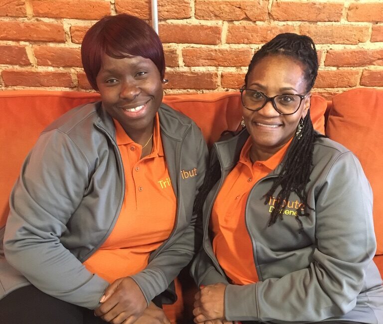 2 Tribute Caregivers smiling together on couch at Tribute Home Care