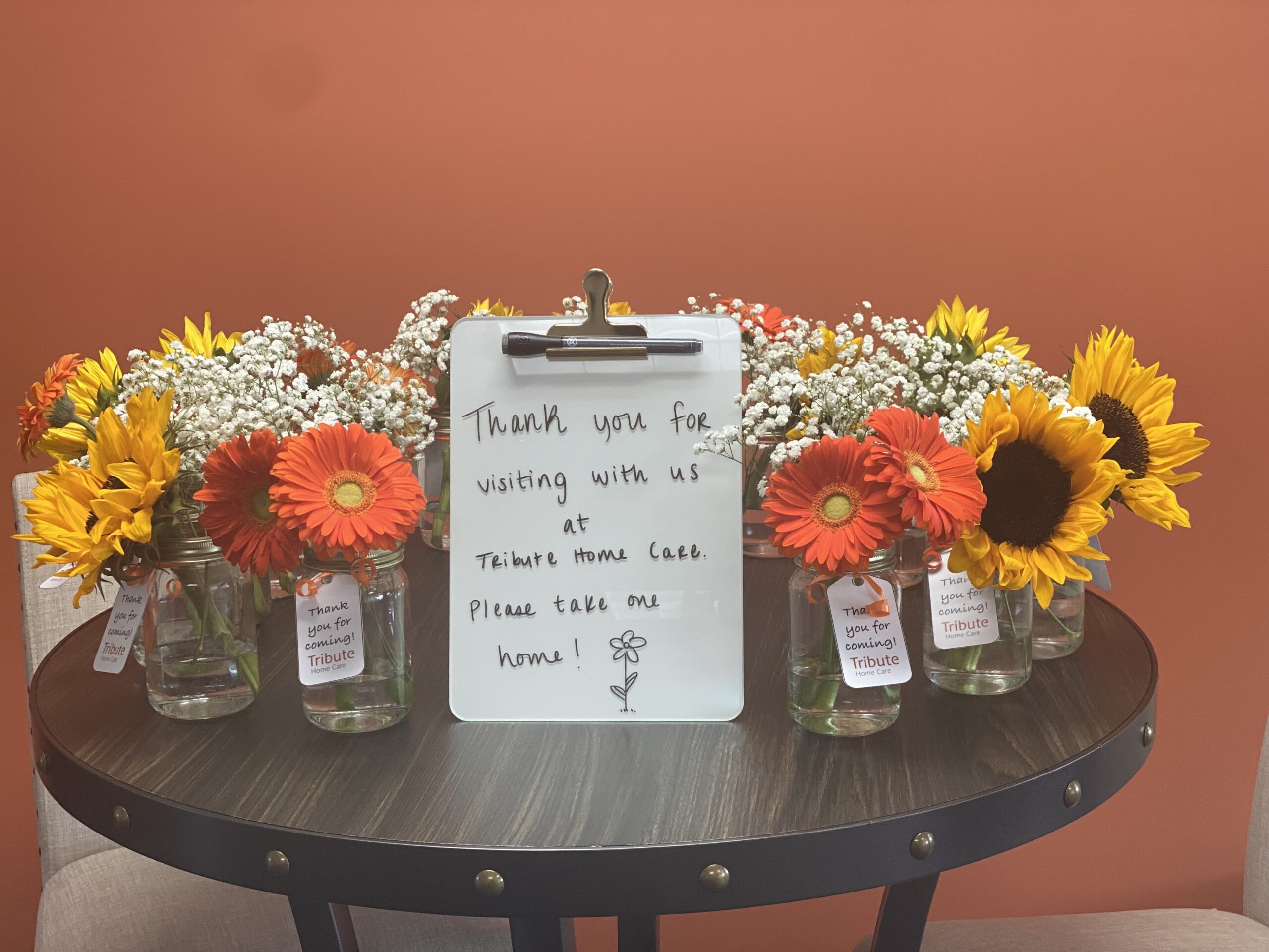 Beautiful flowers in glass mason jars on brown wood table from Tribute. "Thank you for visiting with us at Tribute Home Care. Please take one home!"