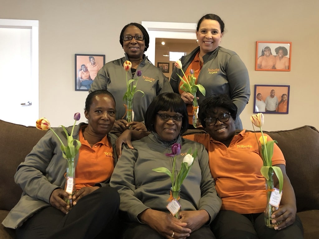 5 Tribute Caregivers smiling and holding flowers together on couch at Tribute Home Care